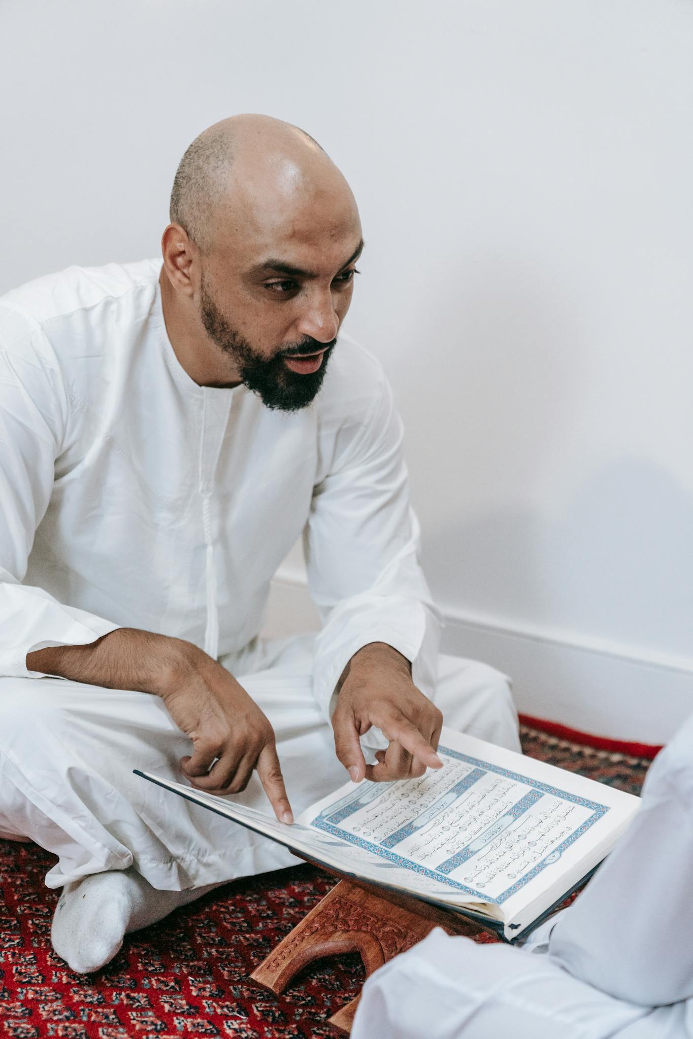 Man sitting indoors in a thobe, reading a religious text from Quran on a wooden stand.